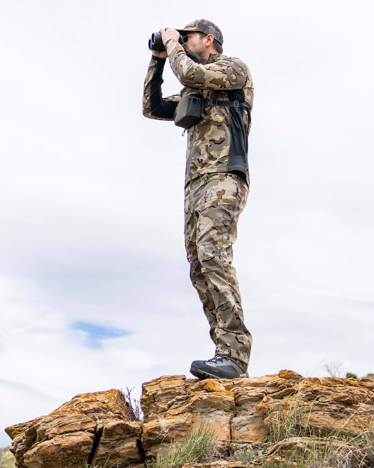 Hunter glassing at the top of a rocky viewpoint, wearing Tiburon Pant in Valo Camouflage