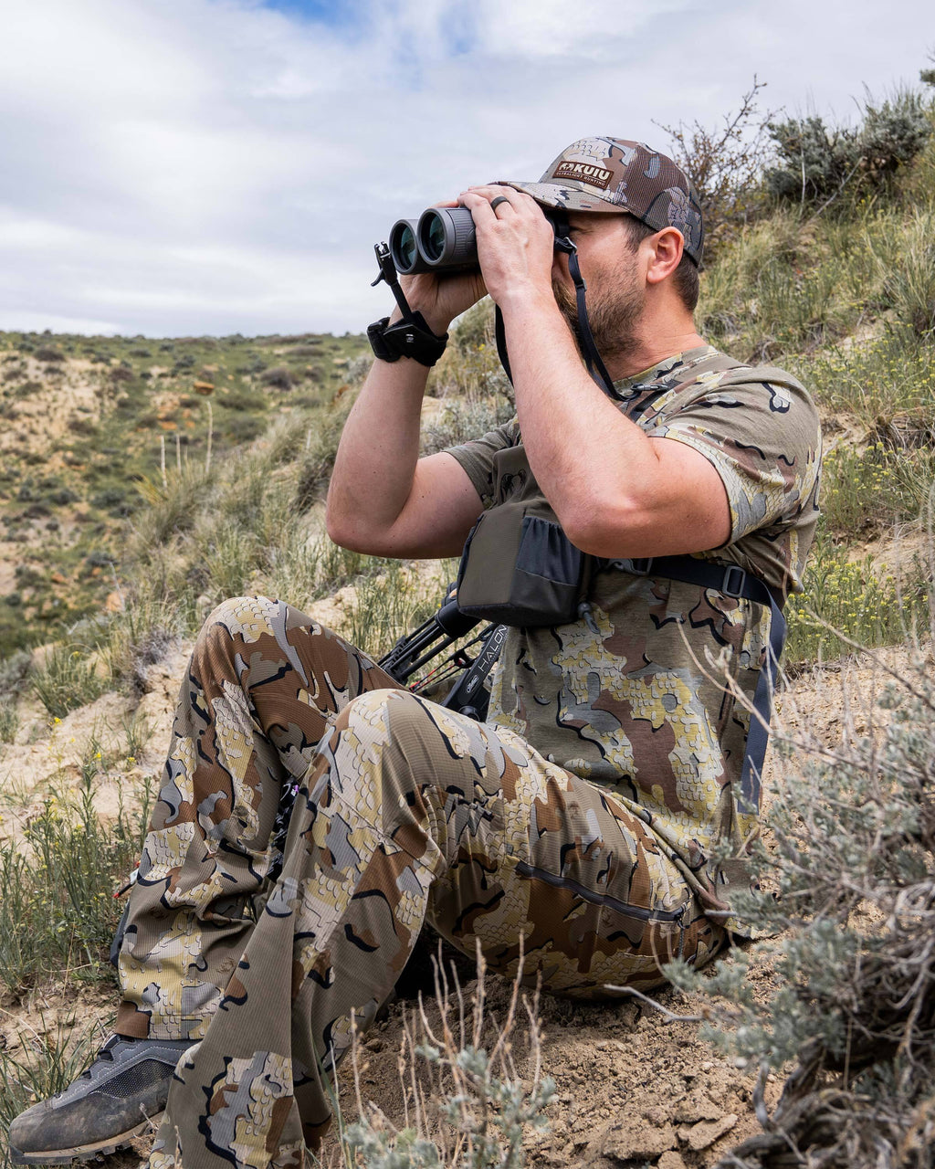 Hunter glassing and sitting on a slopped hillside in the desert, wearing Tiburon Pant in Valo Camouflage