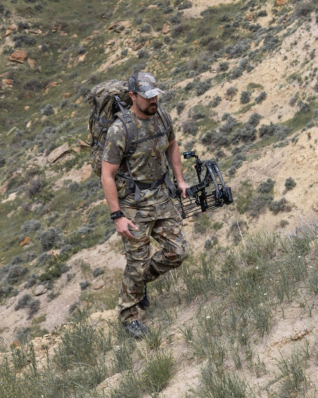 Hunter backpacking up a steep hill, holding a bow in his left hand, wearing Tiburon Pant in Valo Camouflage