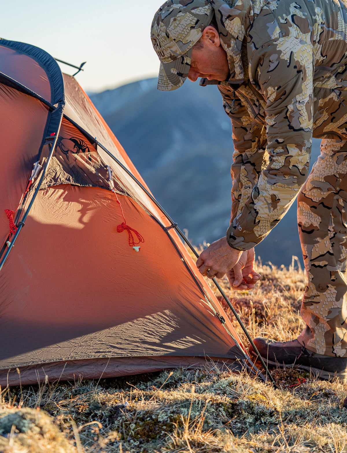 Side of Hunter in Valo Camouflage Adjusting Poles on Mountain Start Two Person Tent in Gunmetal Orange Color