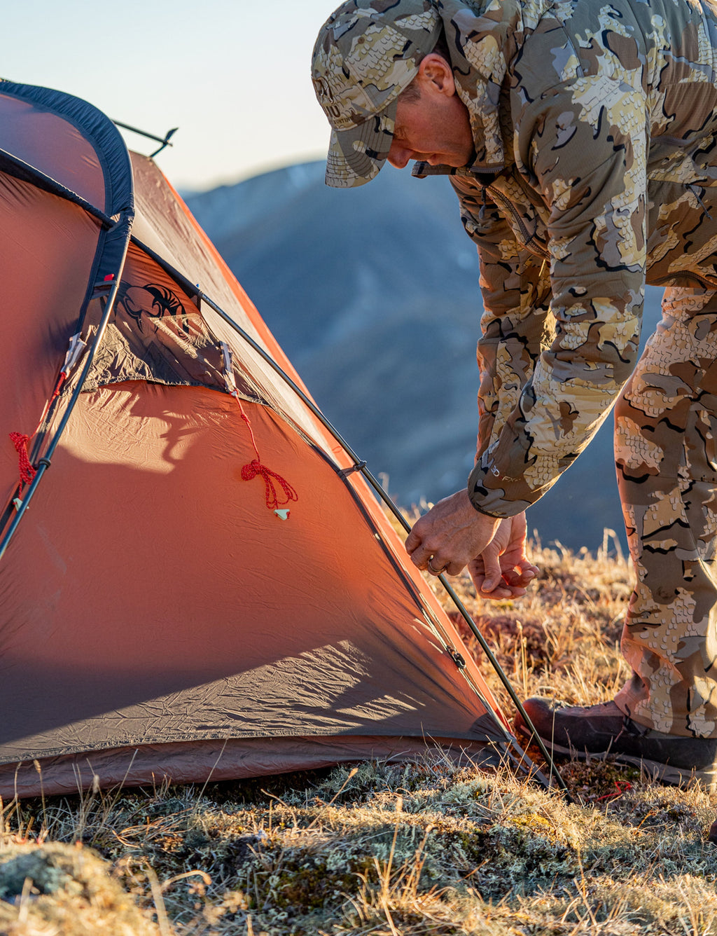 Hunter leans over checking the security of the tent pole on the Mountain Star 2 Person Tent in Gunmetal Grey, Orange