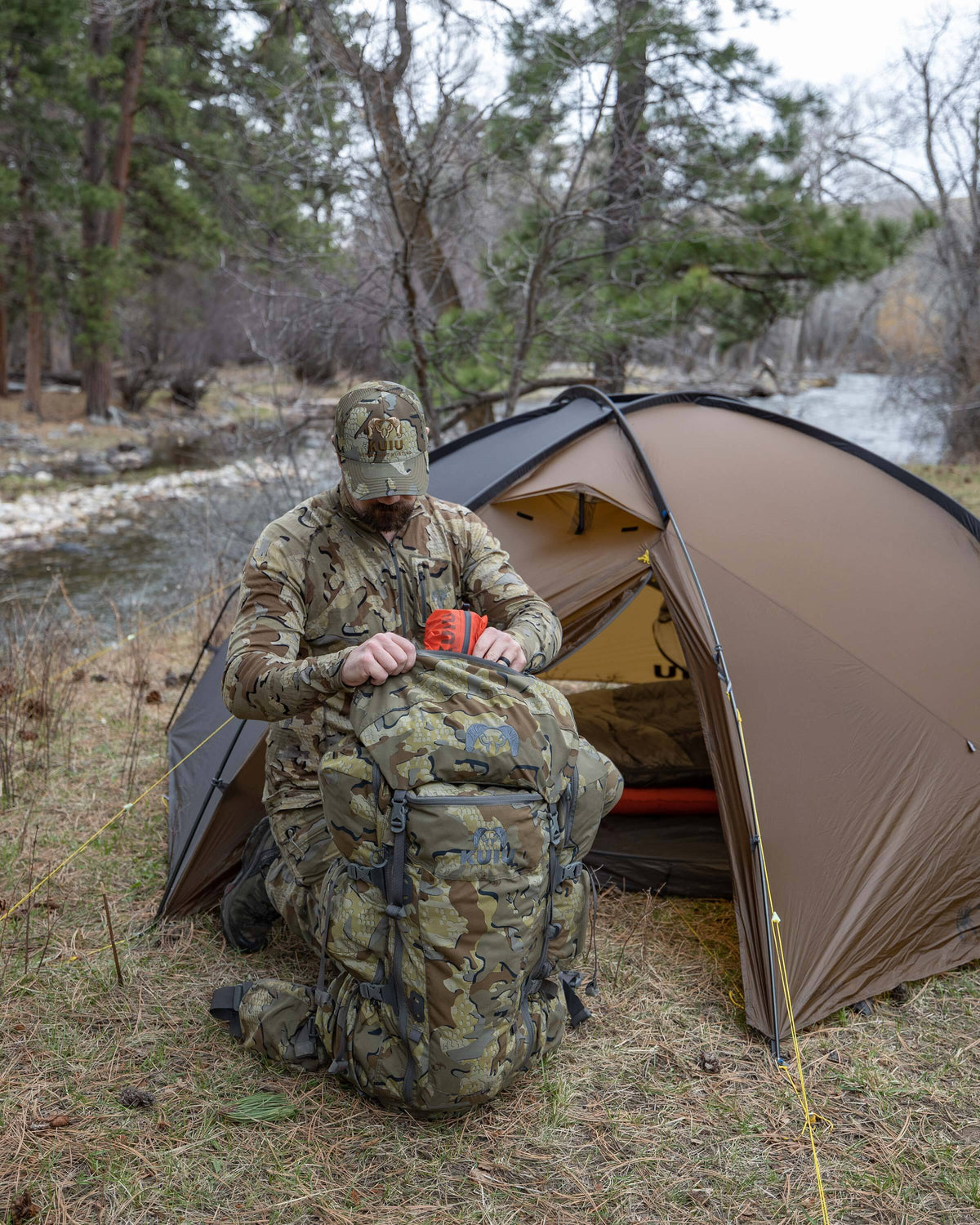 Hunter in Valo Camouflage kneeling in front of a Mountain Star 3 Person Tent in Camel Brown as he loads his PRO LT 7000 Full kit in Valo camouflage