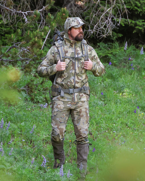 Hunter stands against forest backdrop with both hands on his backpack straps wearing Alpine Hooded Jacket in Valo Camouflage