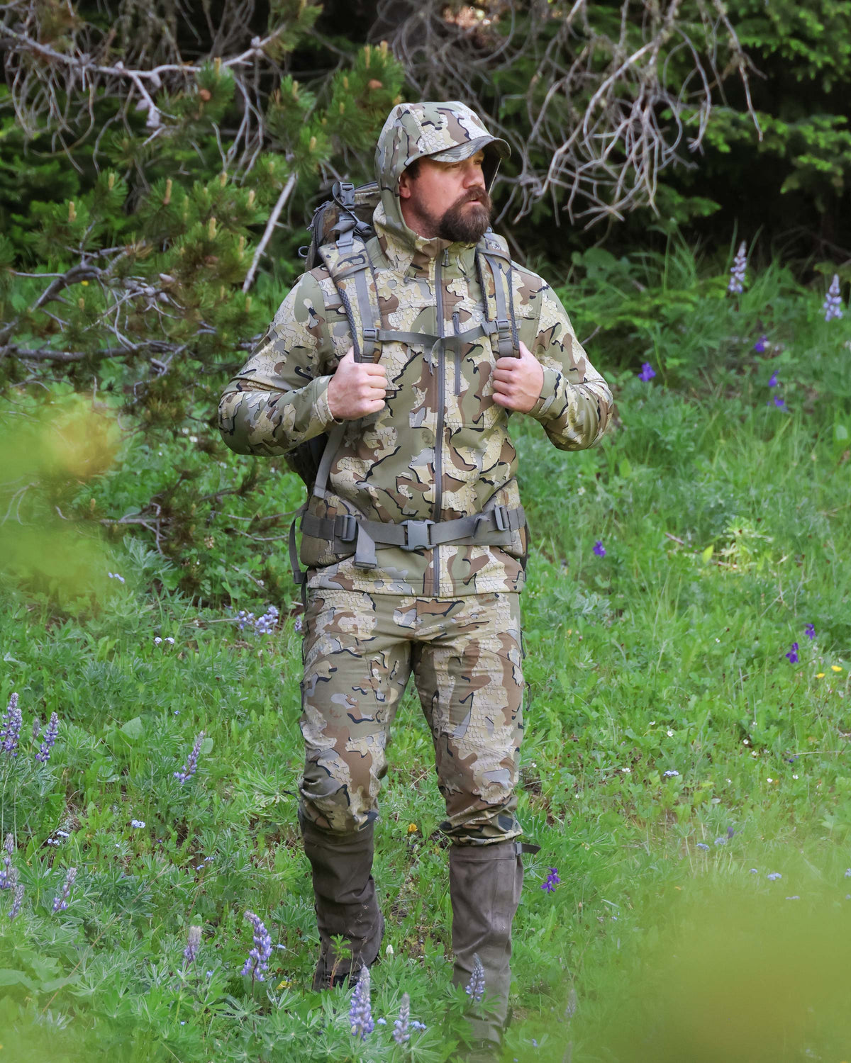 Hunter stands against forest backdrop with both hands on his backpack straps wearing Alpine Hooded Jacket in Valo Camouflage