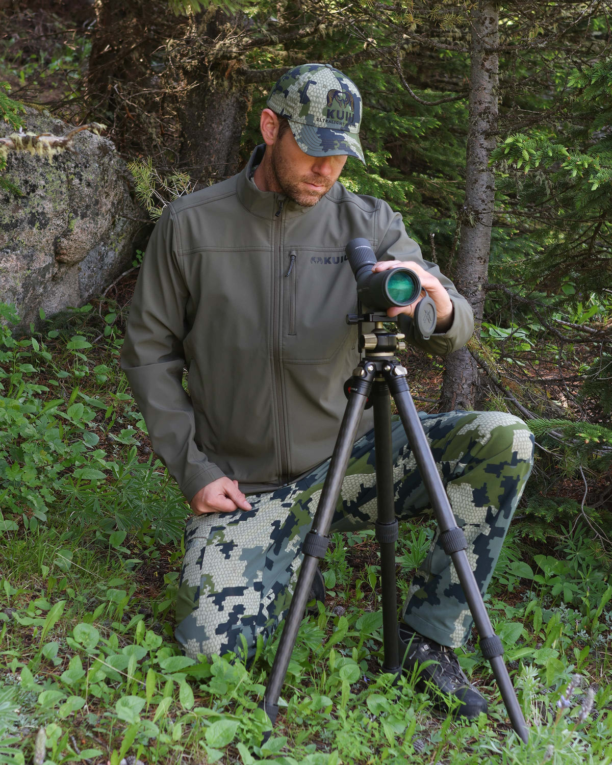 Hunter adjusting in his spotting scope on a tripod wearing Alpine Jacket in Ash Brown