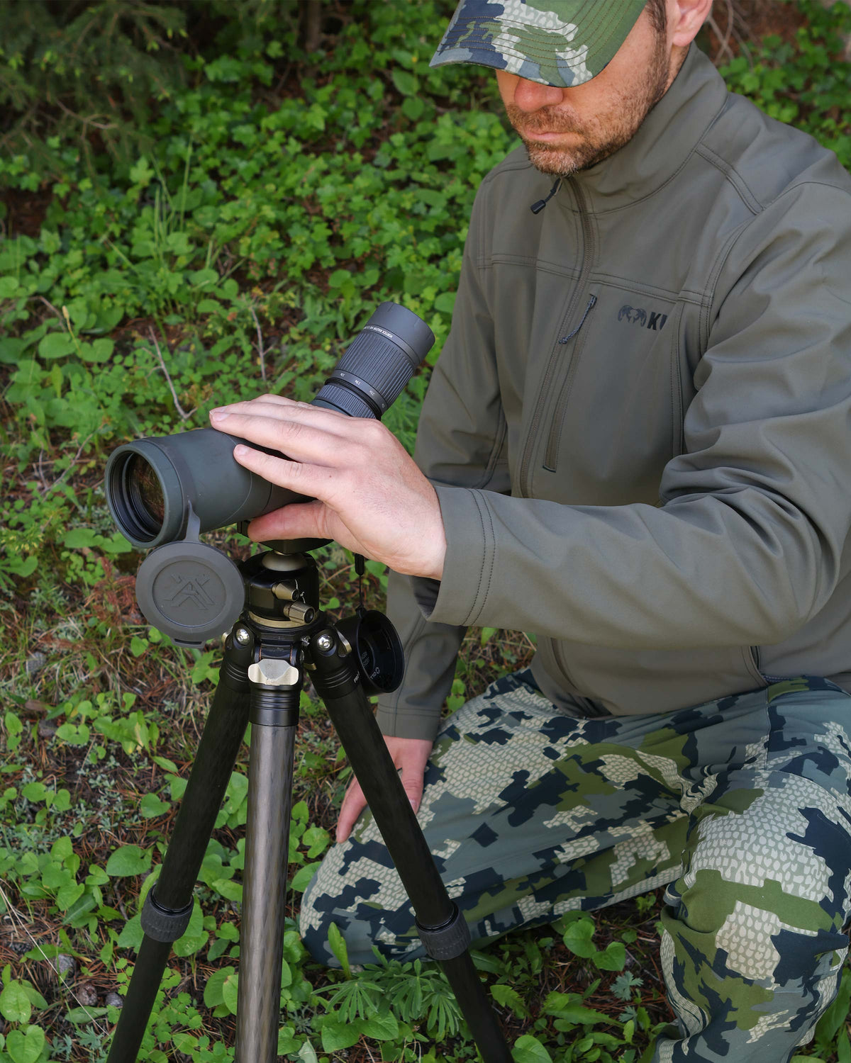 Hunter kneeling down adjusting the spotting scope on a tripod wearing Alpine Jacket in Ash Brown