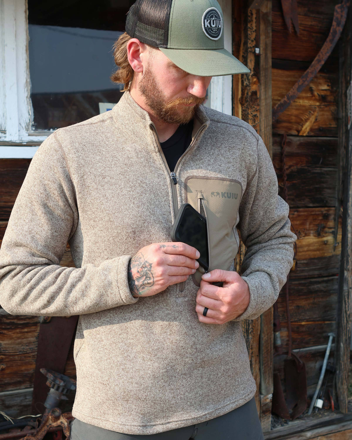 Hunter slipping a cell phone in his left chest pocket, wearing Base Camp Pullover Sweater in Khaki Heather