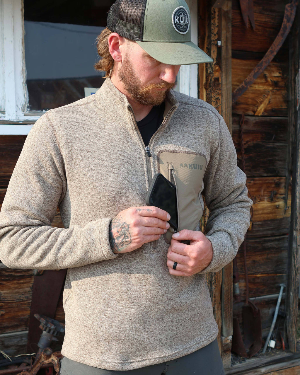 Hunter slipping a cell phone in his left chest pocket, wearing Base Camp Pullover Sweater in Khaki Heather