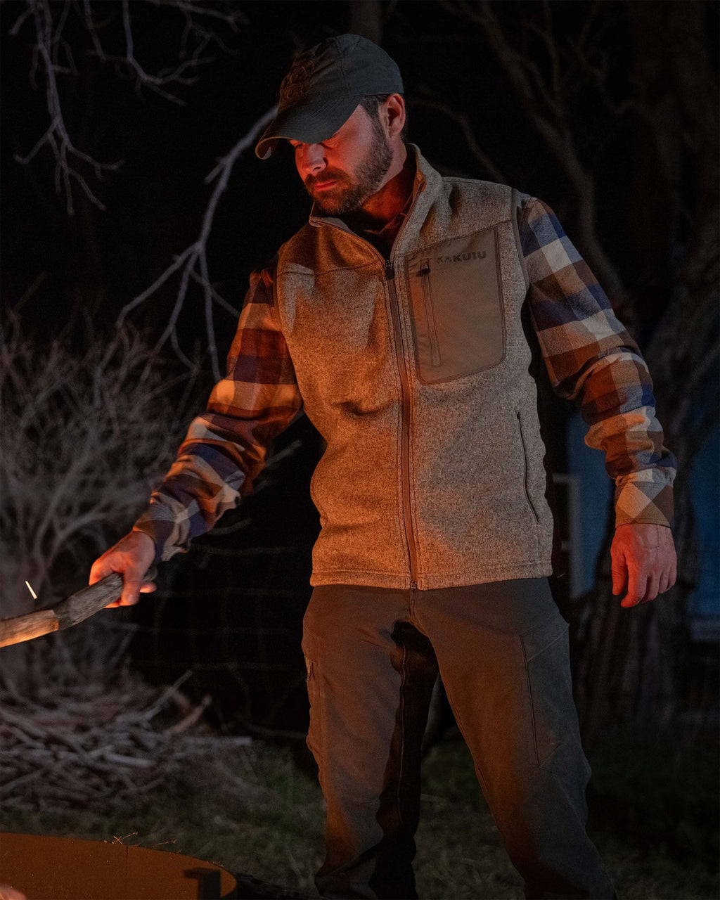 Man in a dark outdoor setting, wearing Base Camp Sweater Vest in Khaki Heather, holding a tree branch over a firepit