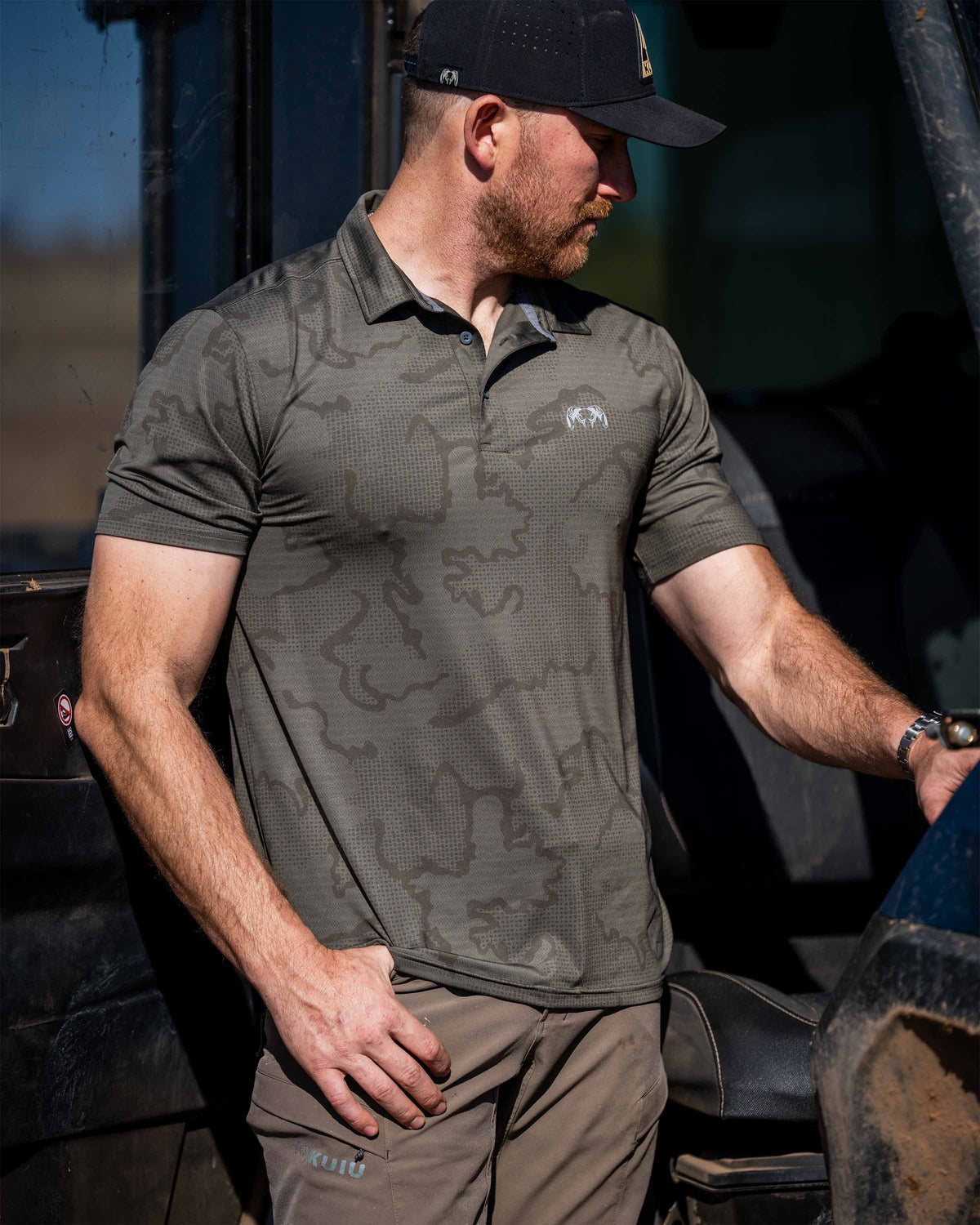 Man is standing next to a working truck, wearing KUIU Performance Polo in Ash Brown Tonal.