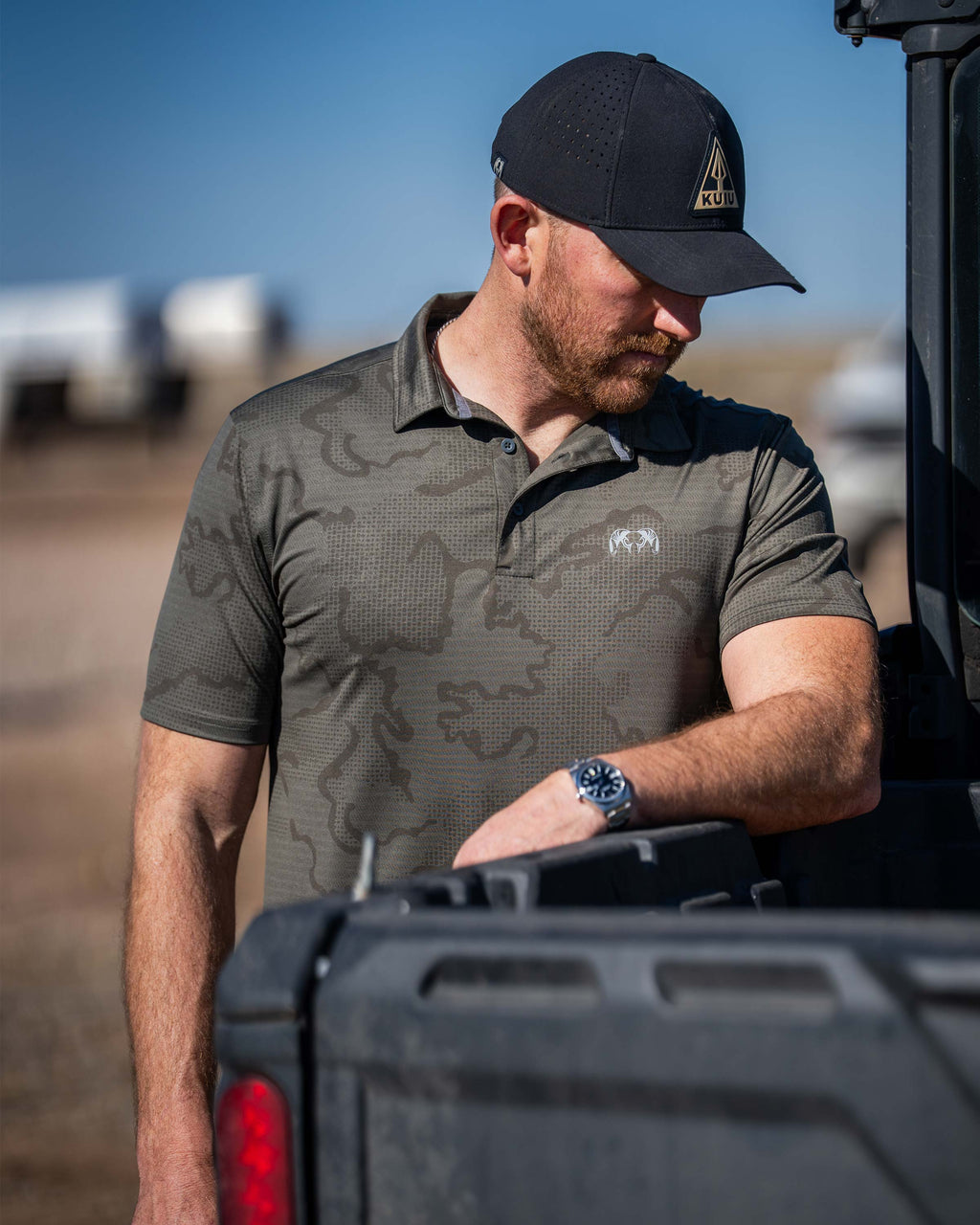 Man is wearing KUIU Performance Polo in Ash Brown Tonal while his arm is resting on the edge of a vehicle.