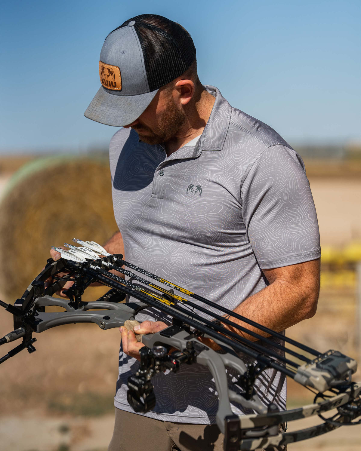 Man is wearing KUIU Performance Polo in Dark Lunar Topo, looking down at a compound bow with multiple arrows loaded.