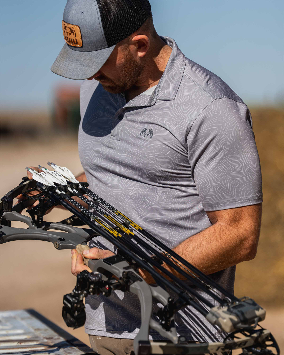 Man is wearing KUIU Performance Polo in Dark Lunar Grey Topo, looking down at a compound bow with multiple arrows loaded.