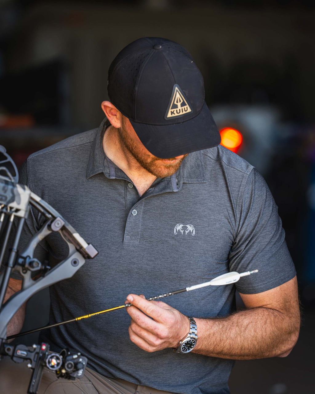 Man stares down at the fletched end of the arrow, wearing KUIU Performance Polo in Heather Stone Grey.
