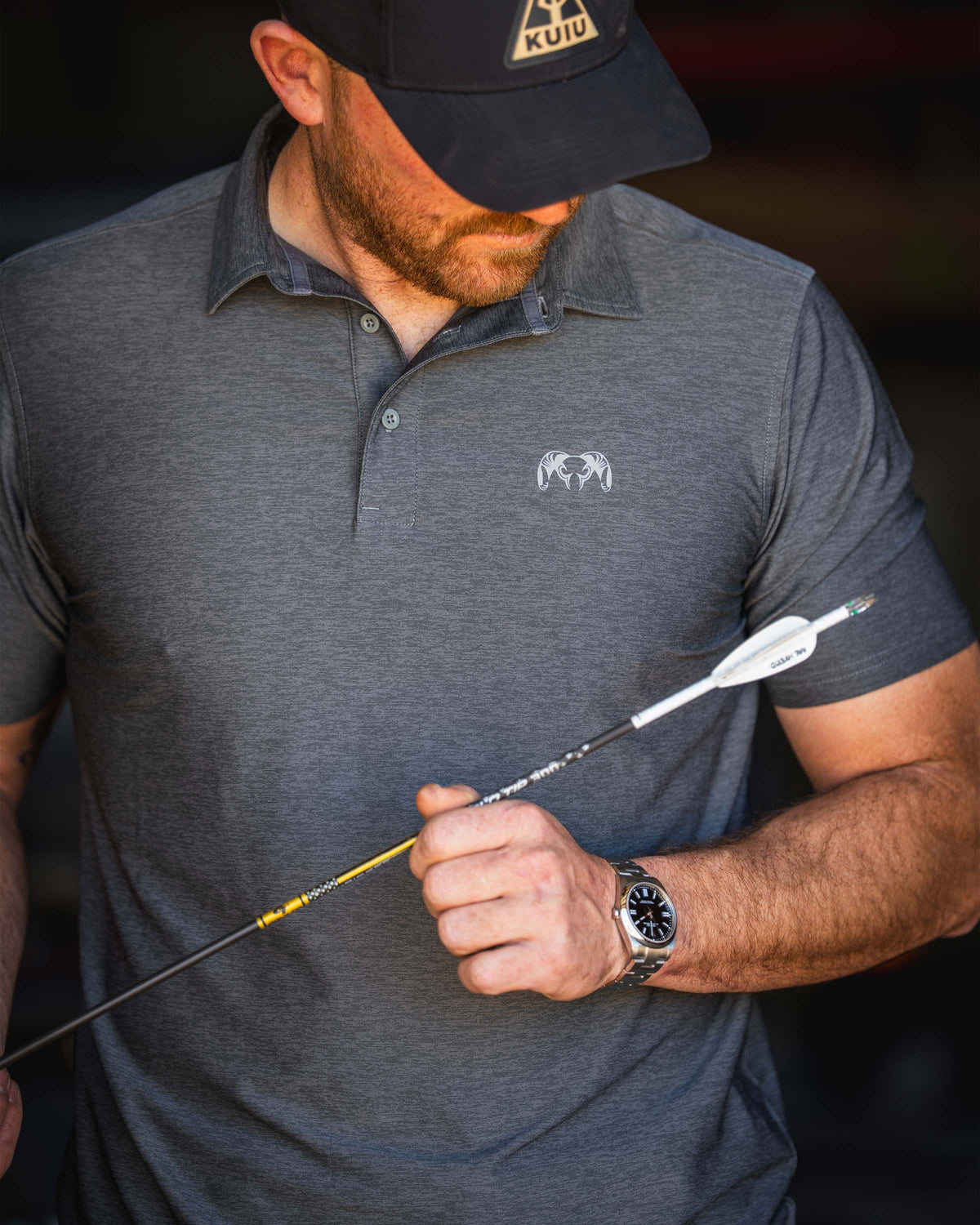 Man stares down at the fletched end of the arrow, wearing KUIU Performance Polo in Heather Stone Grey.