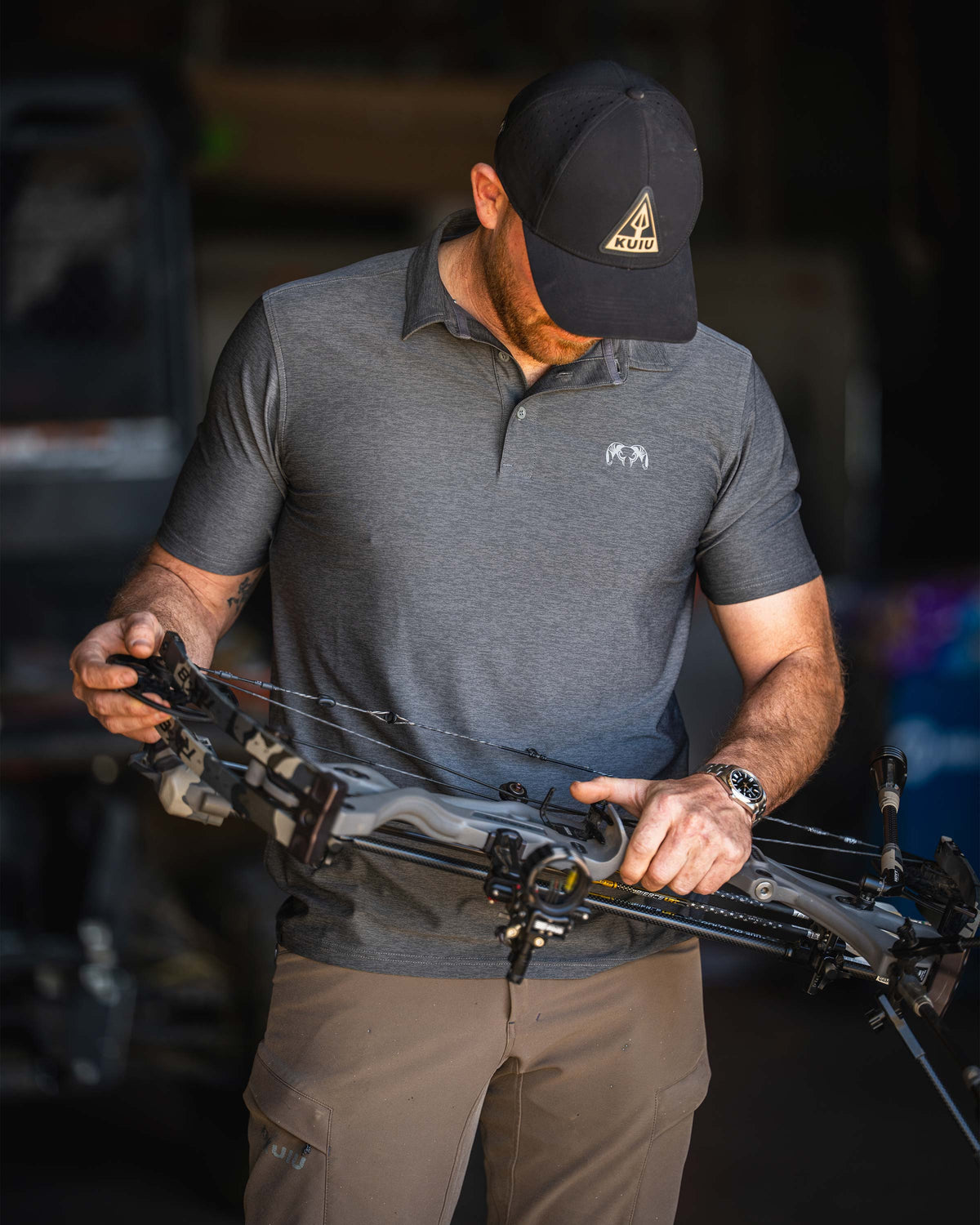 Man inspecting his compound bow, wearing KUIU Performance Polo in Heather Stone Grey.