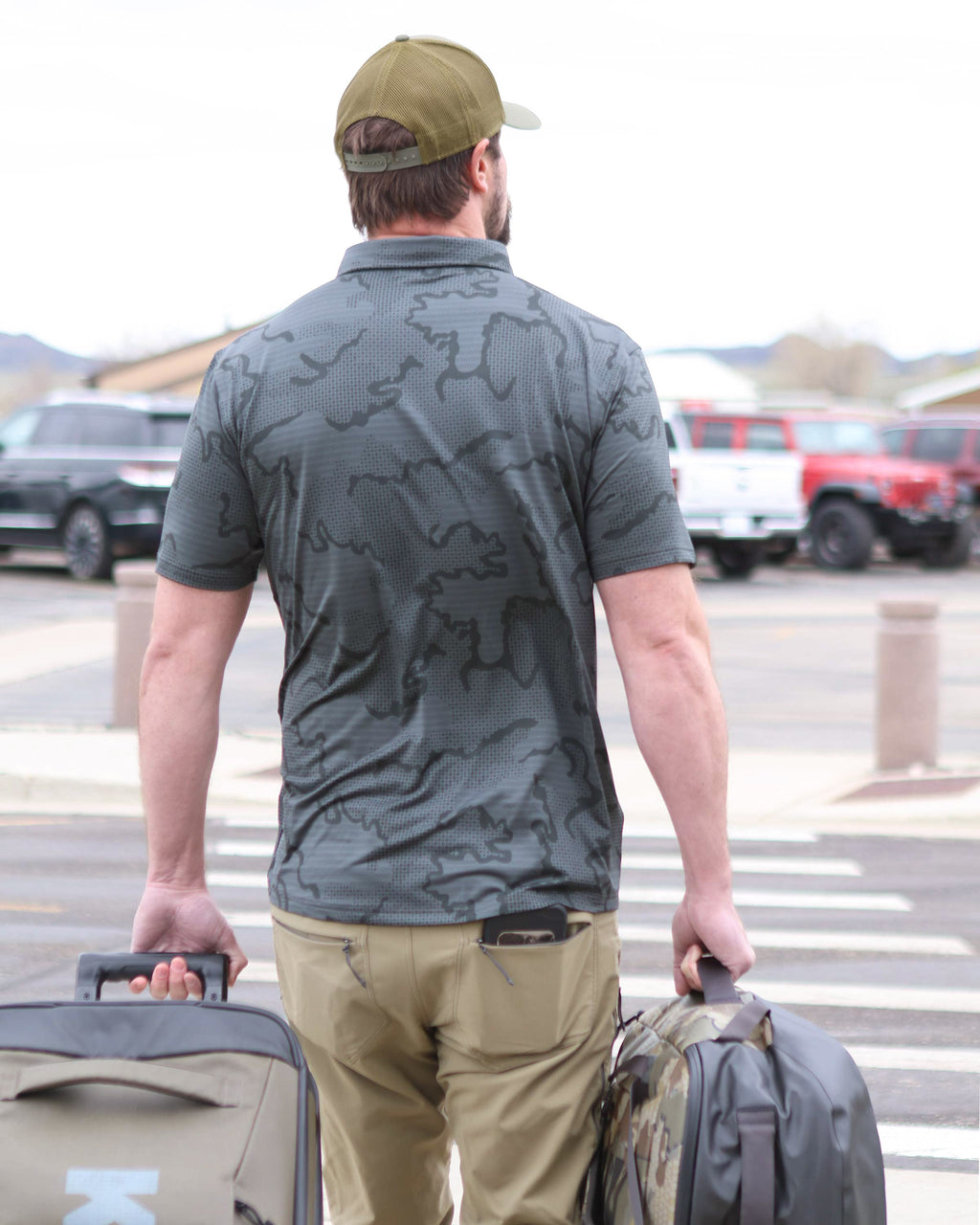 Back view of Hunter walking towards airport parking lot with his luggages, wearing KUIU Performance Polo T-Shirt in Gunmetal Grey Tonal Print