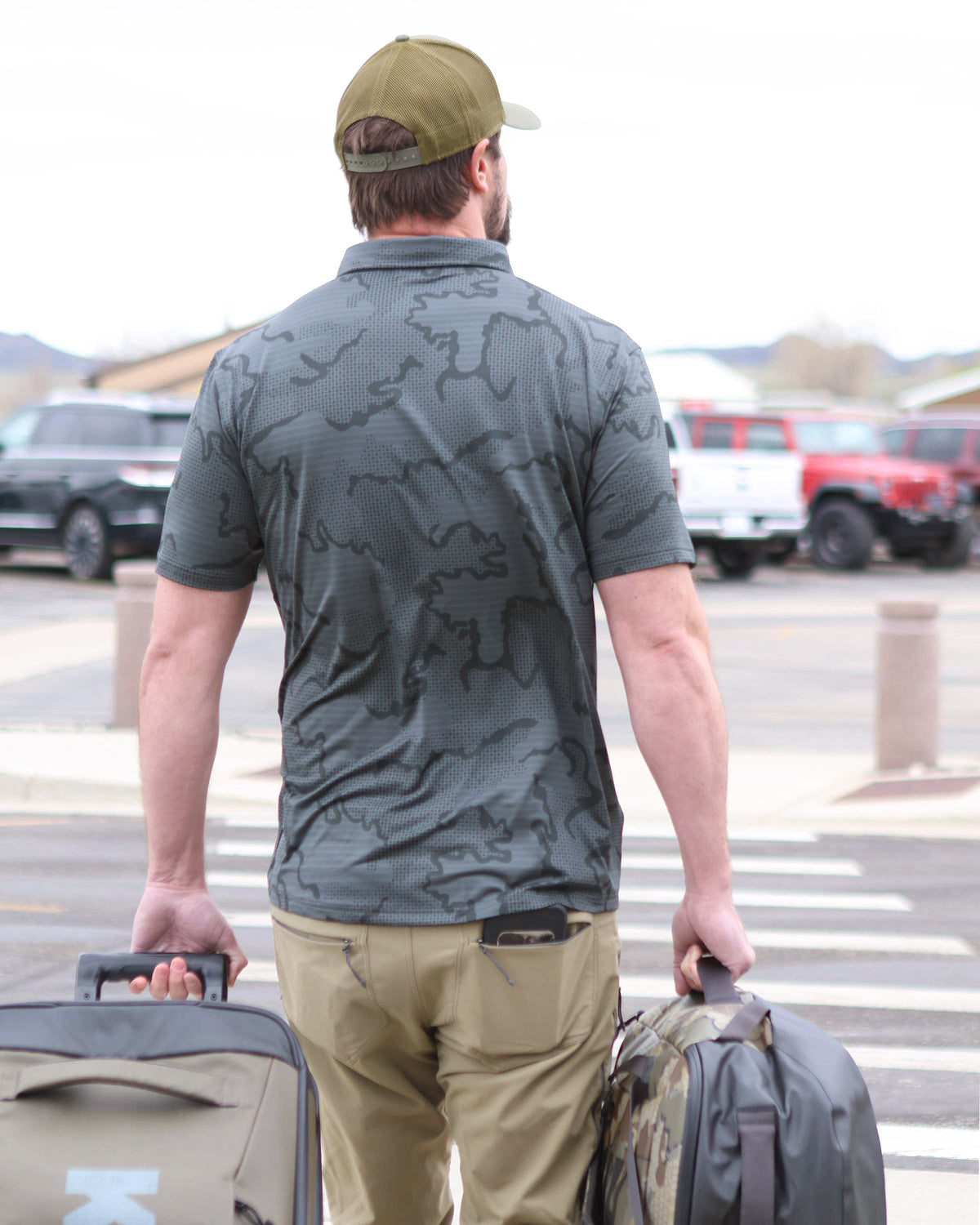 Back view of Hunter walking towards airport parking lot with his luggages, wearing KUIU Performance Polo T-Shirt in Gunmetal Grey Tonal Print