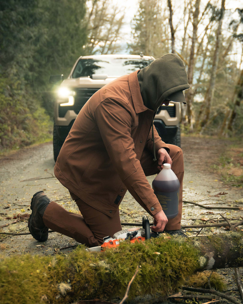 Man kneeling down about to add gas in his chainsaw, wearing Brawley Canvas Jacket in Saddle Brown