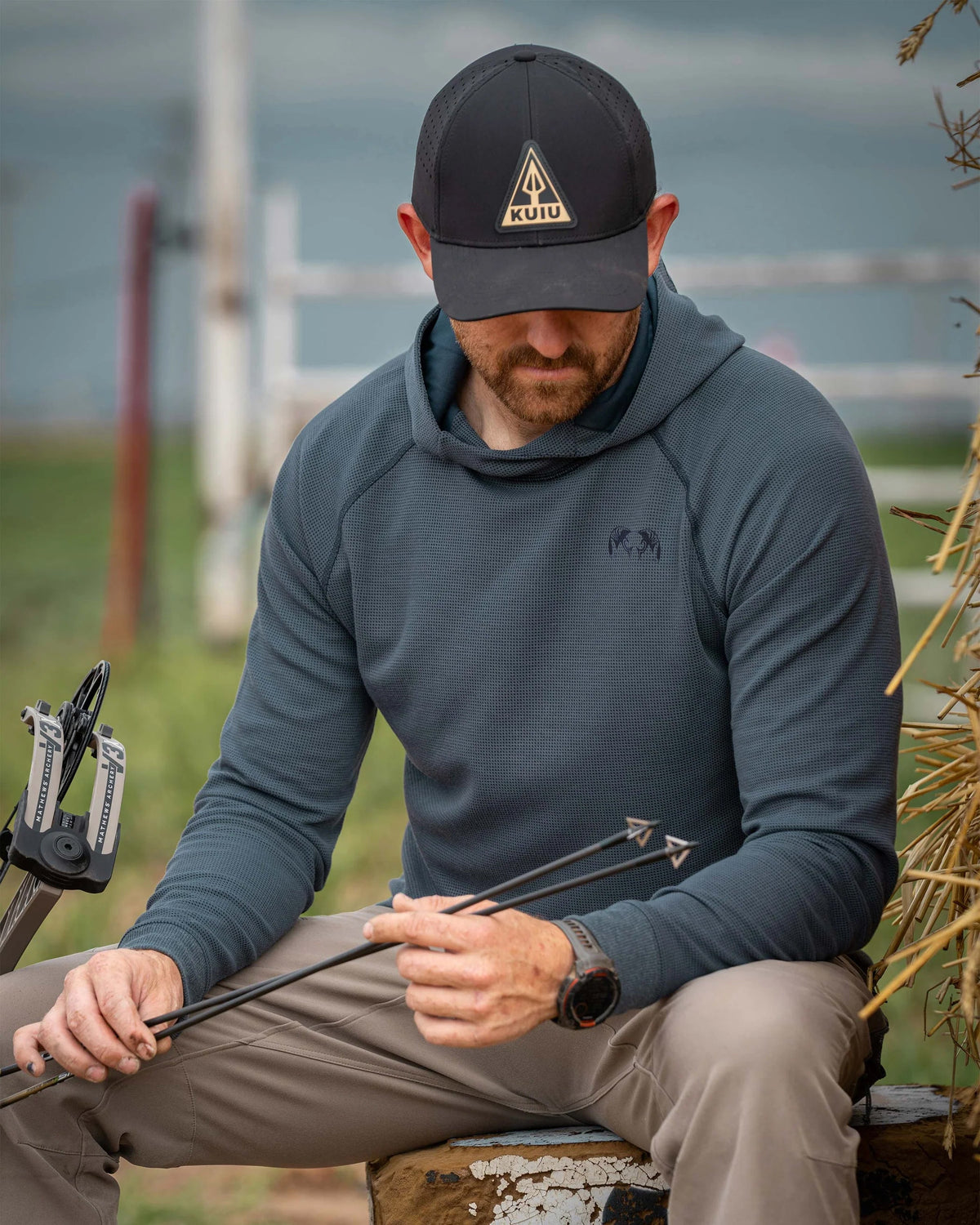 Man sitting on a wooded stump, accessing arrows in his hand, wearing Canyon Waffle Hoodie in Steel Blue