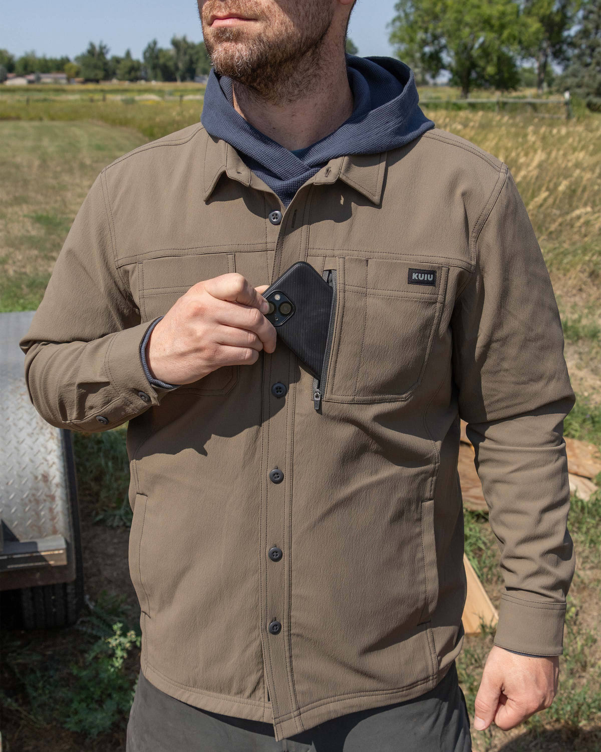 Man in an outdoor setting wearing Motive Flannel Lined Shirt Jacket in Major Brown, putting a cell phone in the left vertical zipper pocket