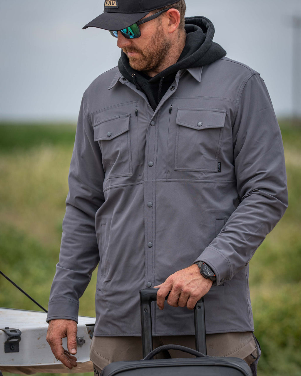 Man wearing Tour Shirt Jacket in Castle Rock Grey standing behind a Waypoint Roller Bag