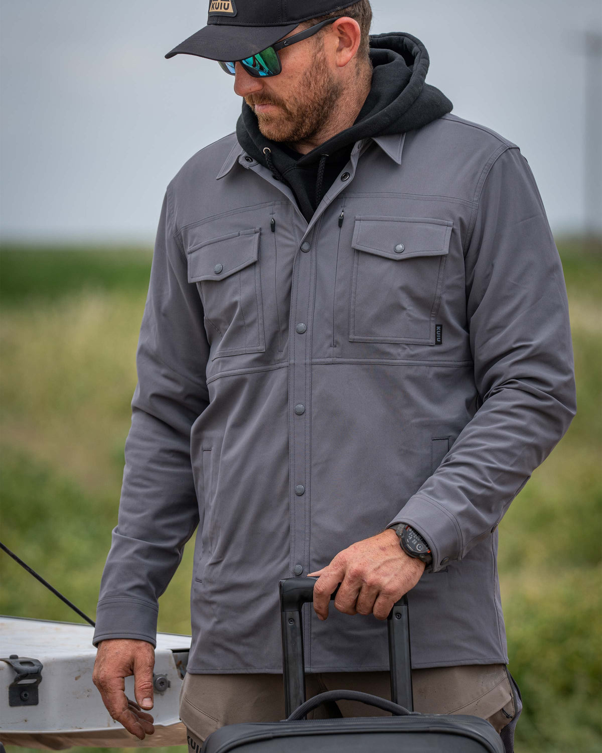 Man wearing Tour Shirt Jacket in Castle Rock Grey standing behind a Waypoint Roller Bag