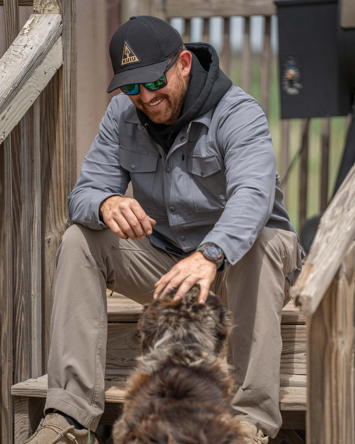 Man smiling sitting on a wooden deck steps petting a dog wearing Tour Shirt Jacket in Castle Rock Grey