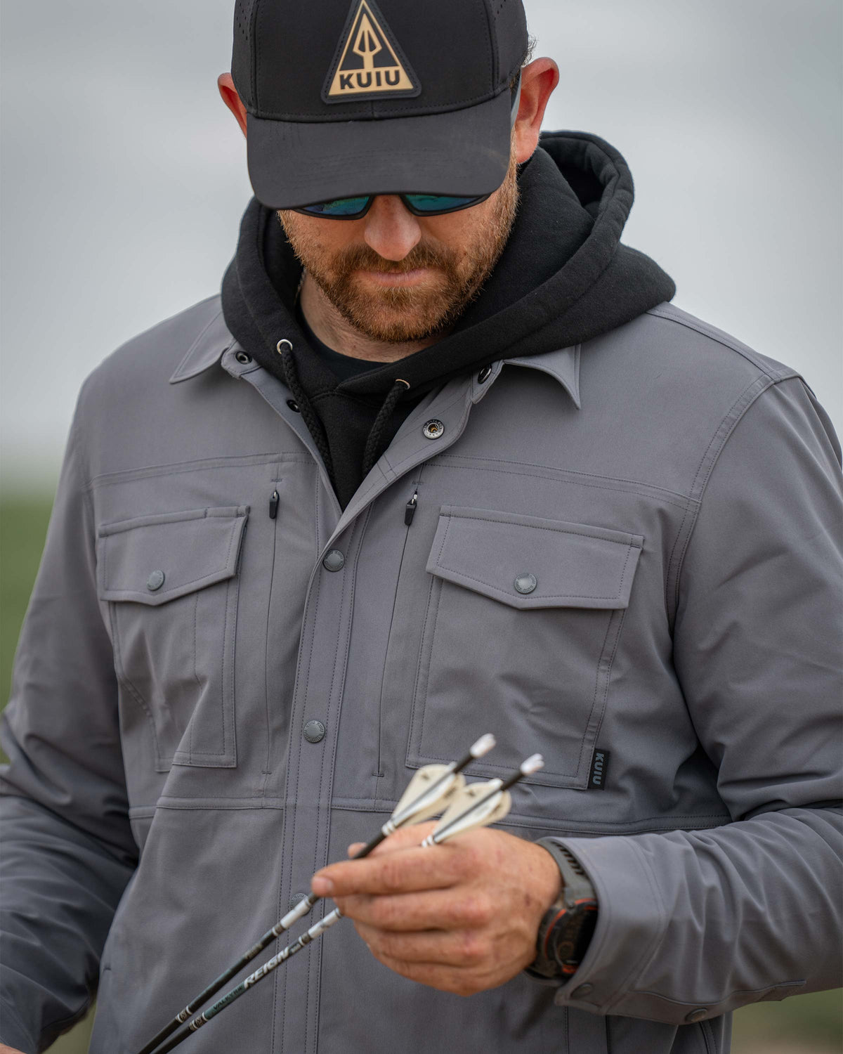 Man examines the fletching part of the arrows he's holding wearing Tour Shirt Jacket in Castle Jacket Grey