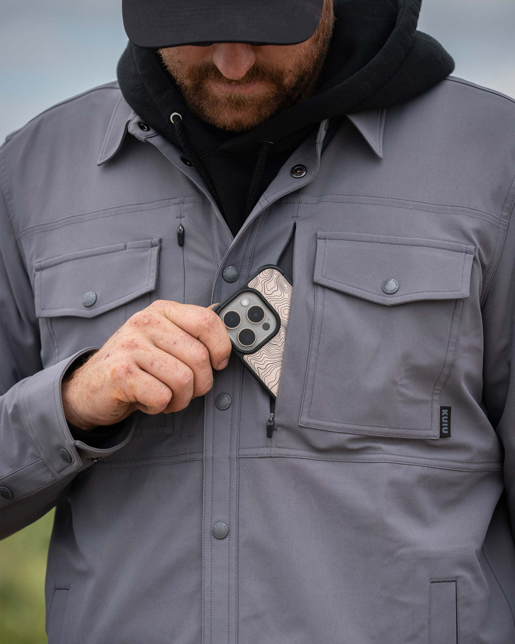 Close-up of man putting a cell phone in the front zippered pocket of the Tour Shirt Jacket in Castle Rock Grey