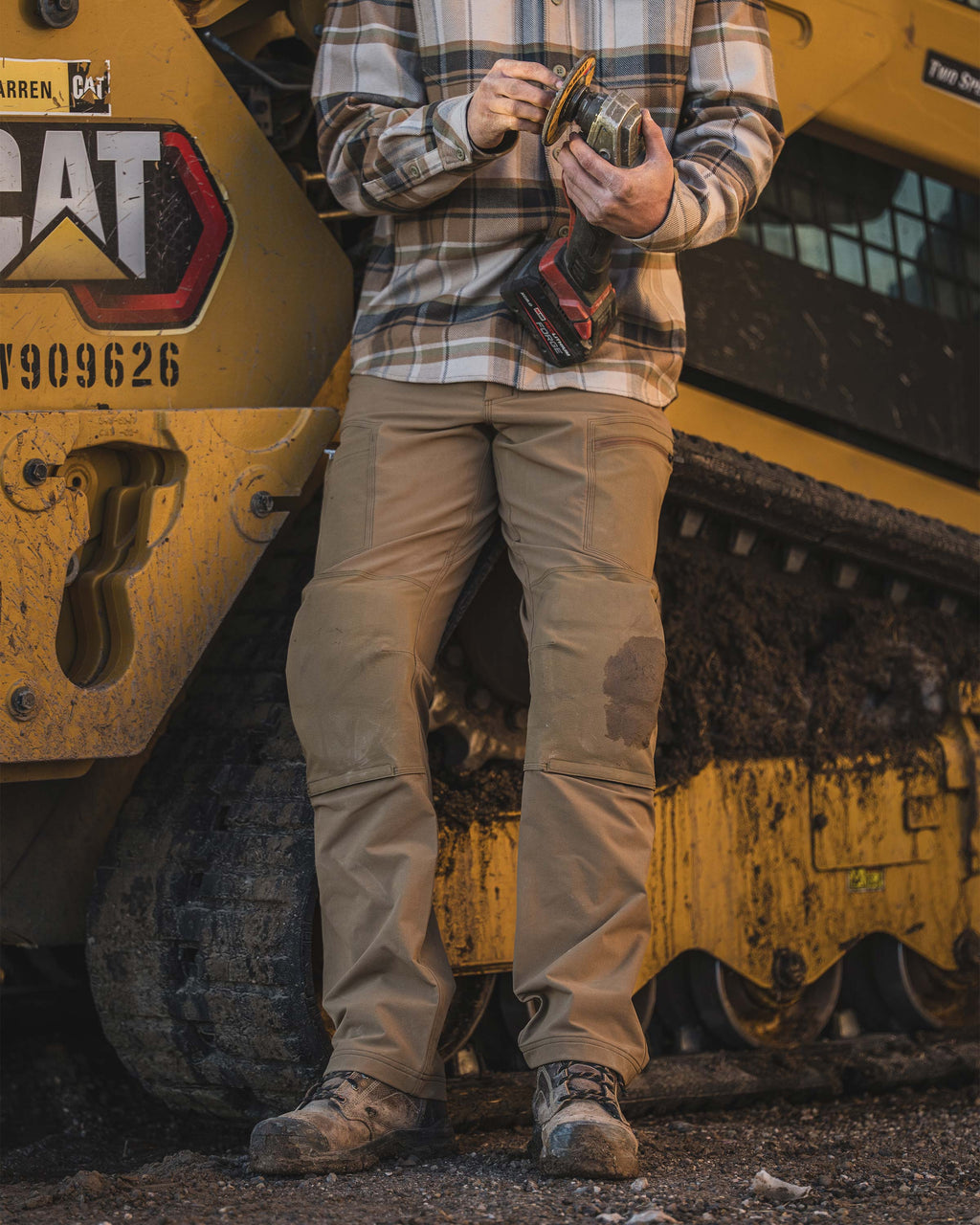 Man leaning against a large yellow Cat brand tracked construction machine, holding a handheld power tool, wearing Groundwork Stretch Woven Pant in Tobacco Brown