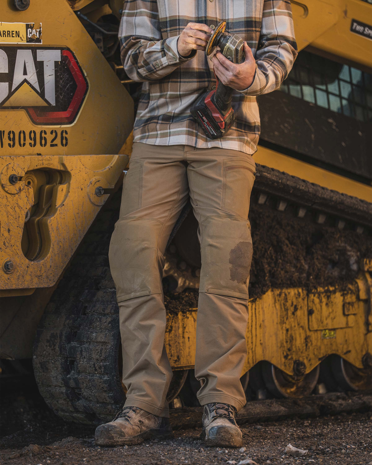Man leaning against a large yellow Cat brand tracked construction machine, holding a handheld power tool, wearing Groundwork Stretch Woven Pant in Tobacco Brown