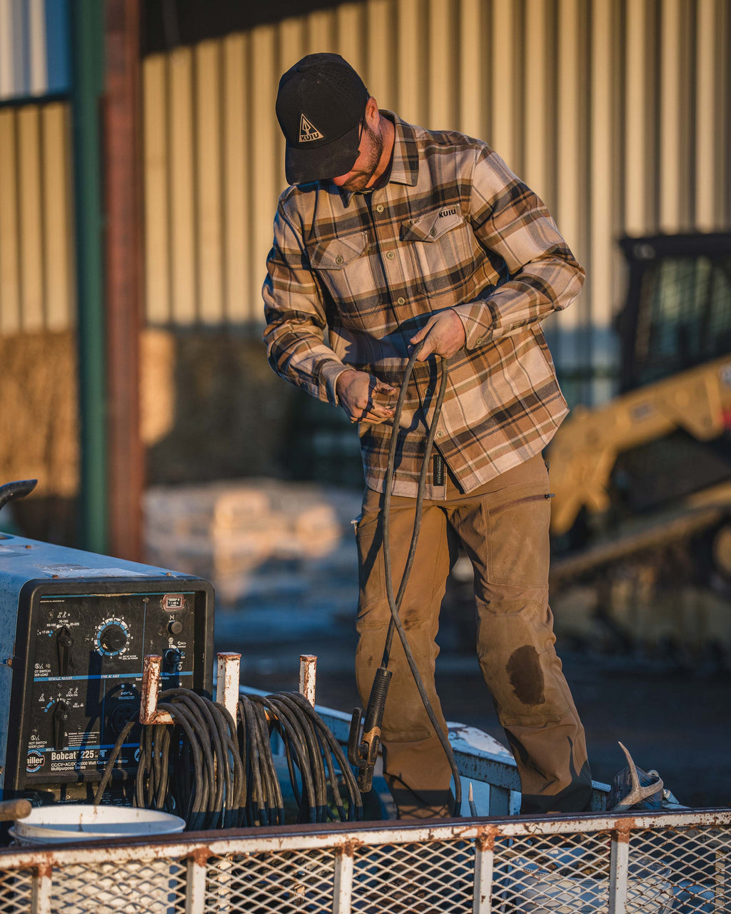 Man standing in a carry-on trailer, Holding several cables appearing to arrange them, Wearing Groundwork Stretch Woven Pant in Tobacco Brown