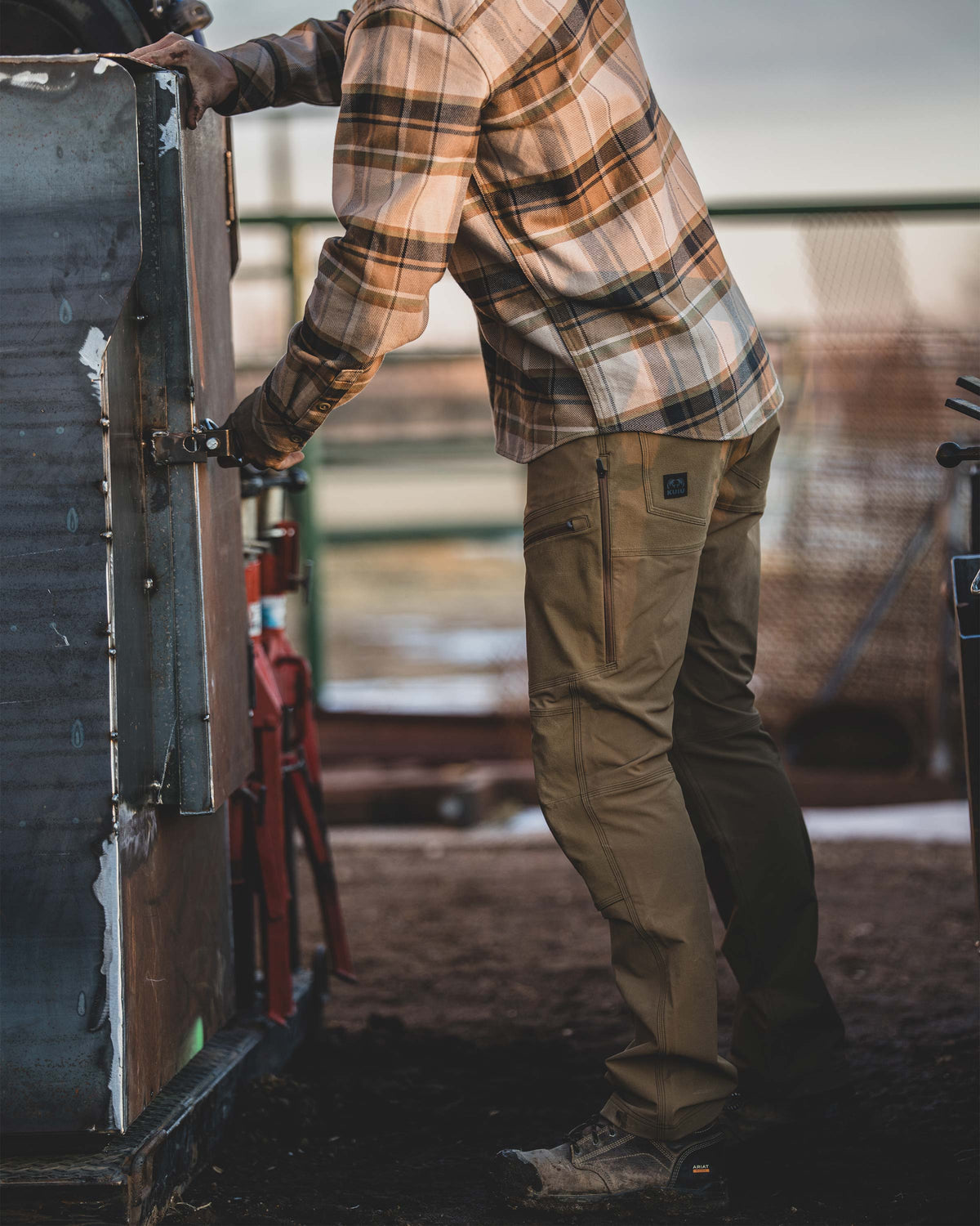 Man standing next to a weathered metal structure adjusting a latch, Wearing Groundwork Stretch Woven Pant in Tobacco Brown