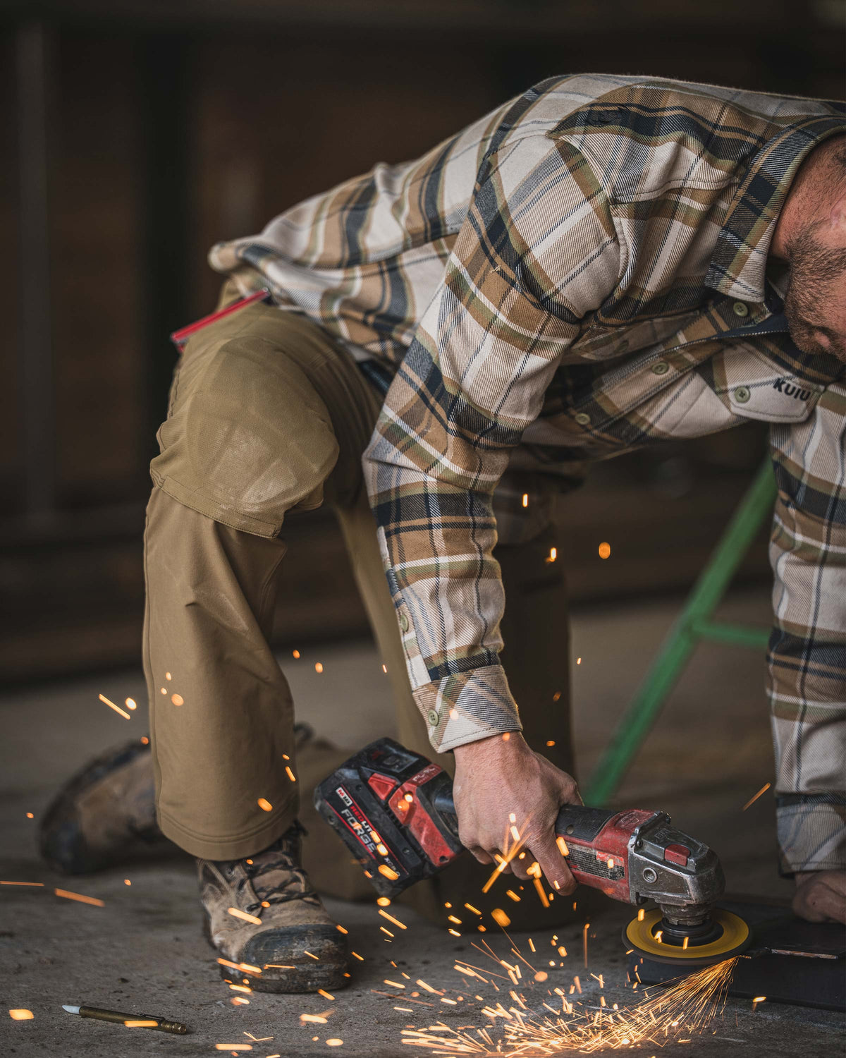 Man wearing Groundwork Stretch Woven Pant in Tobacco Brown, Kneeling on a concrete surface while using a handheld power tool that produces bright sparks as it grinds against a metal object