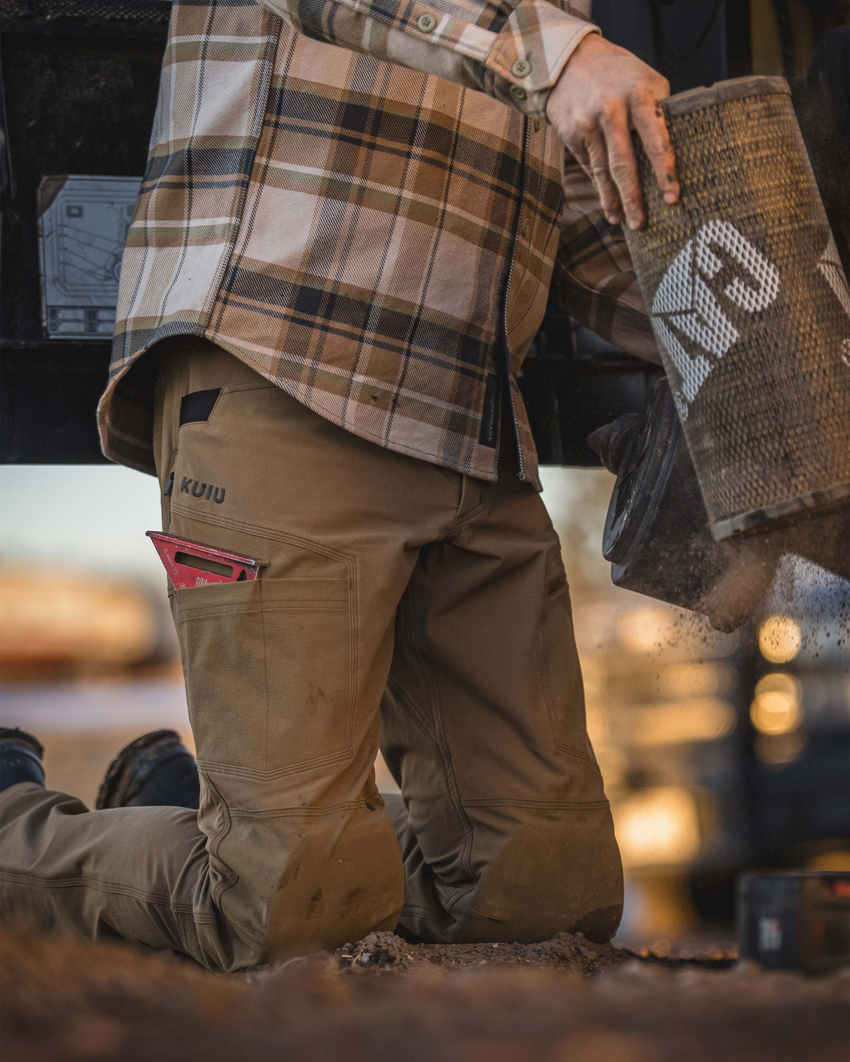 Man kneeling on the ground in an outdoor, work‑site setting, Holding a large cylindrical filter releasing dust as it is handled, Wearing Groundwork Stretch Woven Pant in Tobacco Brown