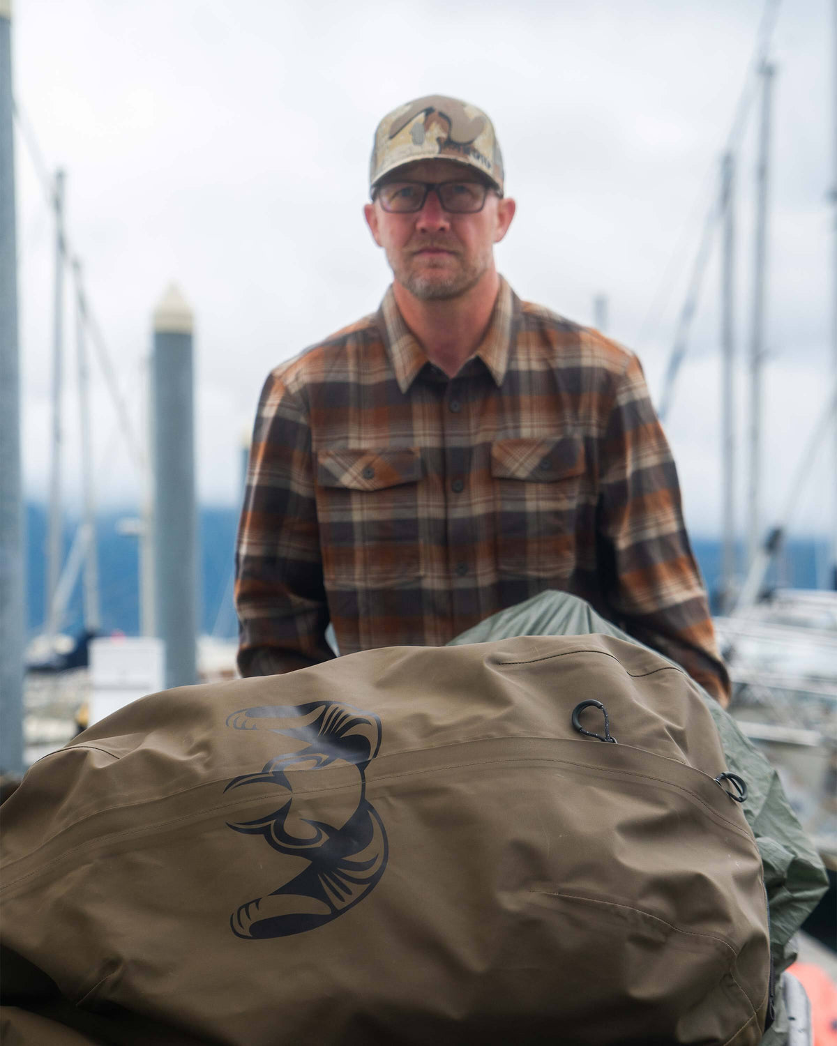 Man leaning in front of the Taku BLSTX 10000 Duffel in Bourbon Brown at a dockside setting, wearing the Field Flannel Shirt in Saddle Blockline.