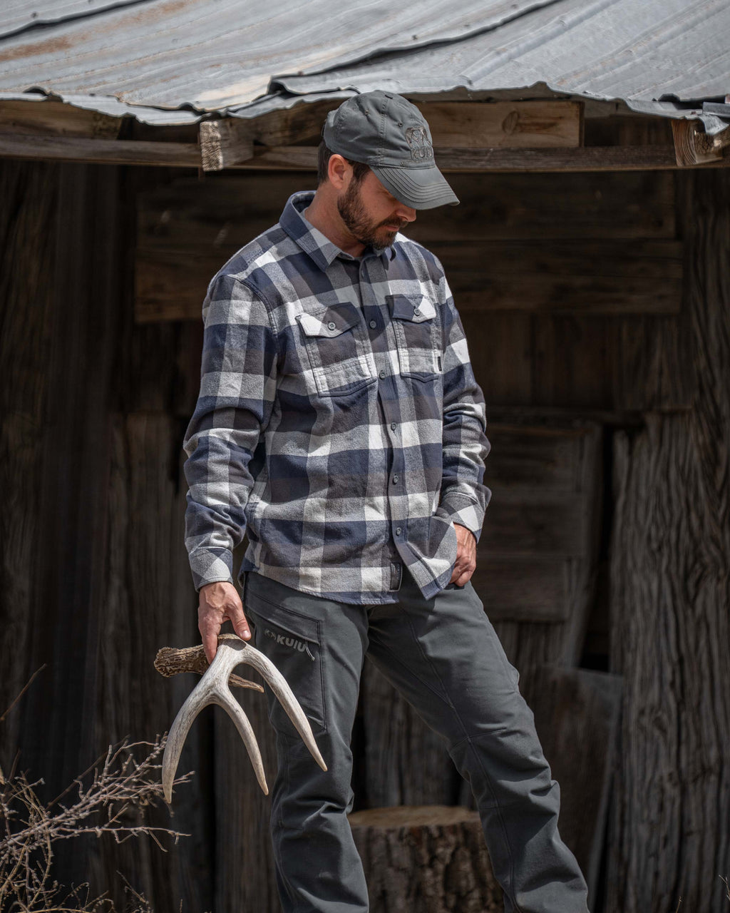 Hunter posing next to a wood cabin, holding a deer antler, wearing Field Flannel Shirt in Steel Blue Trio