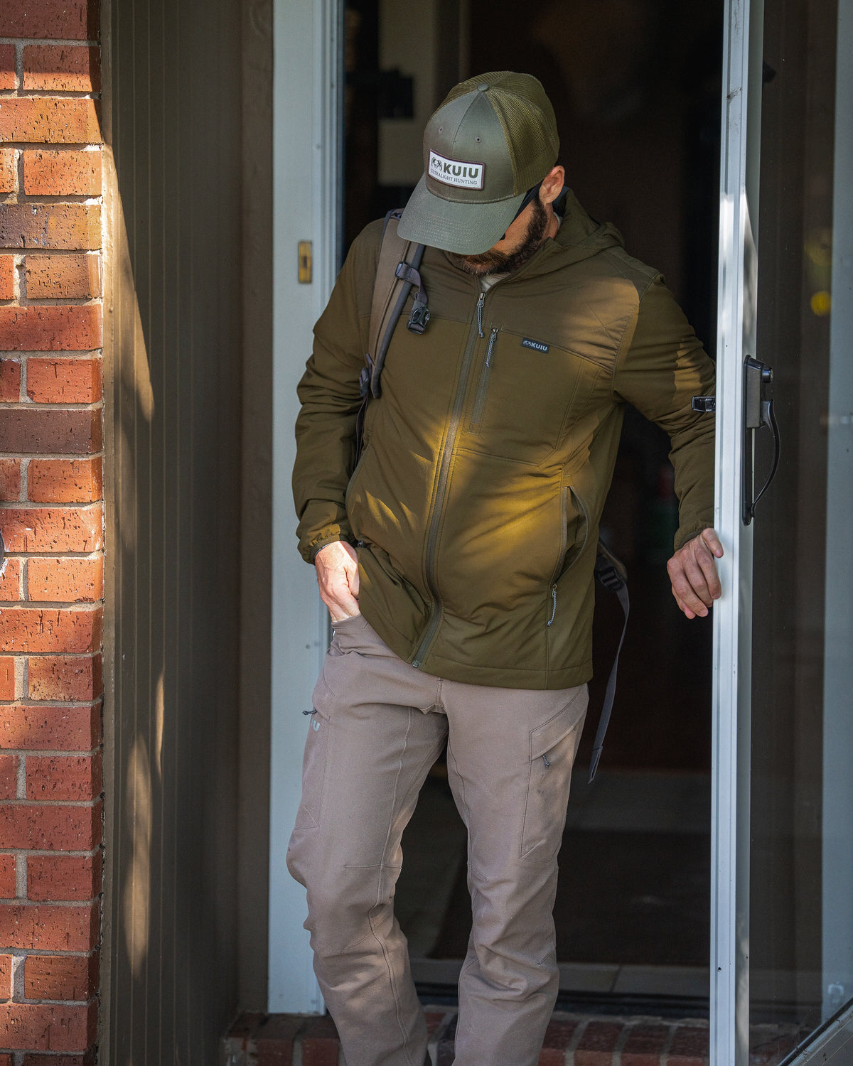 A man wearing Transit Packable Hooded Jacket in Dark Army walks out of a doorway next to a brick wall.