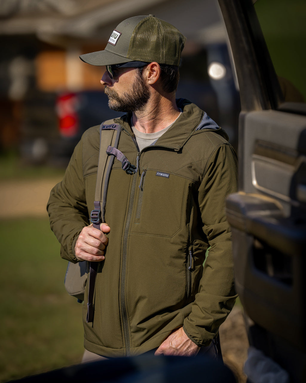 A man wearing a Transit Packable Hooded Jacket in Dark Army/Army Green, standing outdoors next to a vehicle.