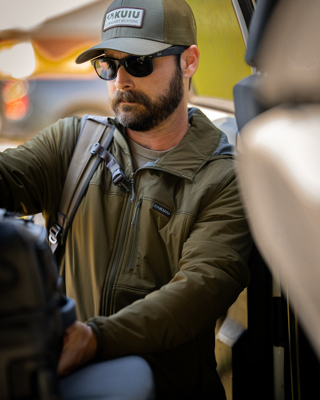 A man wearing Transit Packable Hooded Jacket in Dark Army, sitting inside a vehicle with a backpack.visible.