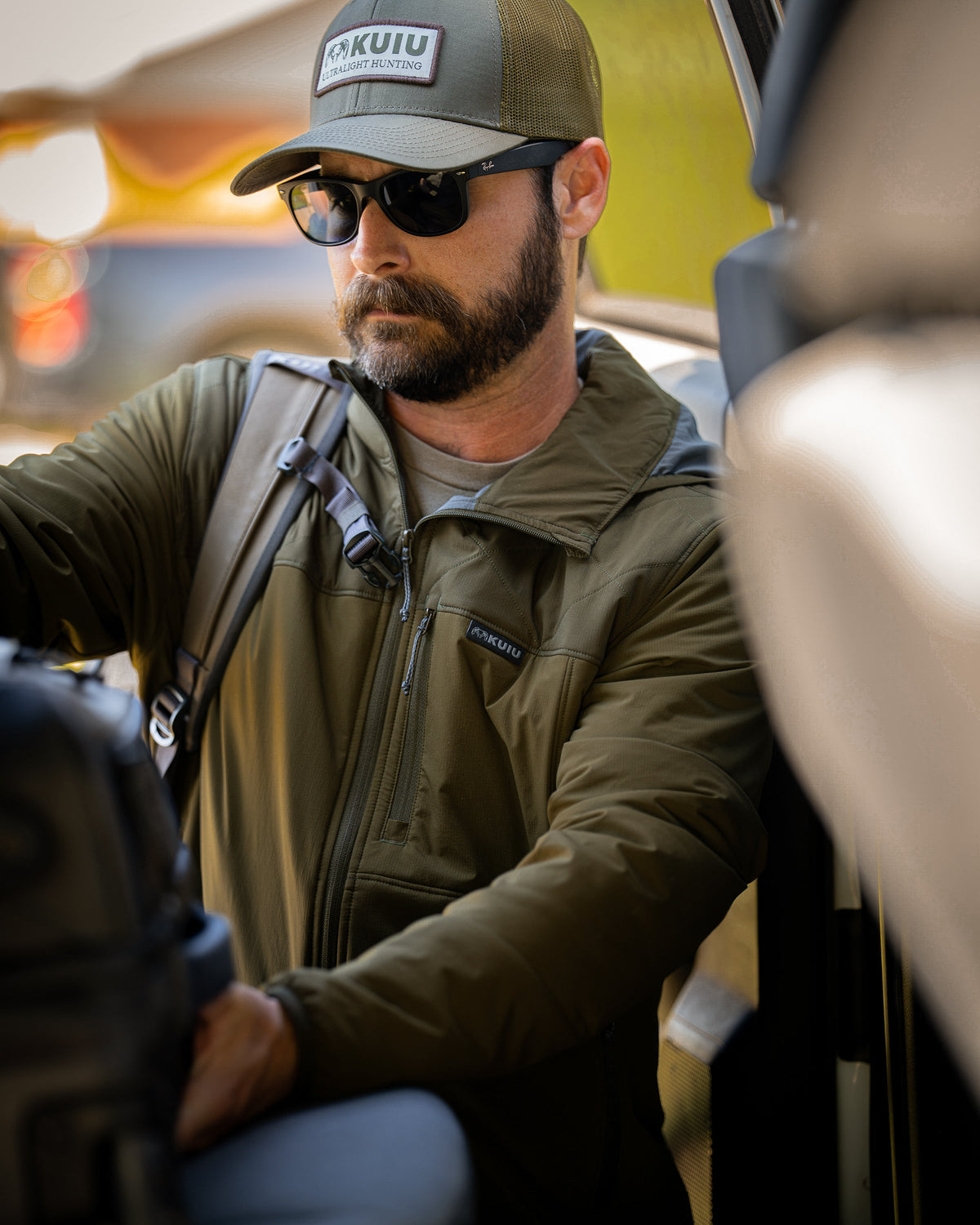 A man wearing Transit Packable Hooded Jacket in Dark Army, sitting inside a vehicle with a backpack.visible.