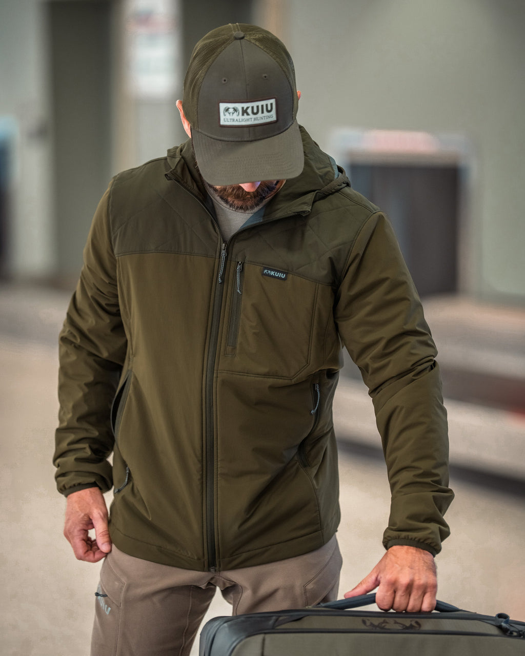 A man wearing a Transit Packable Hooded Jacket in Dark Army/Army Green, standing in a transit area and holding the handle of a suitcase.