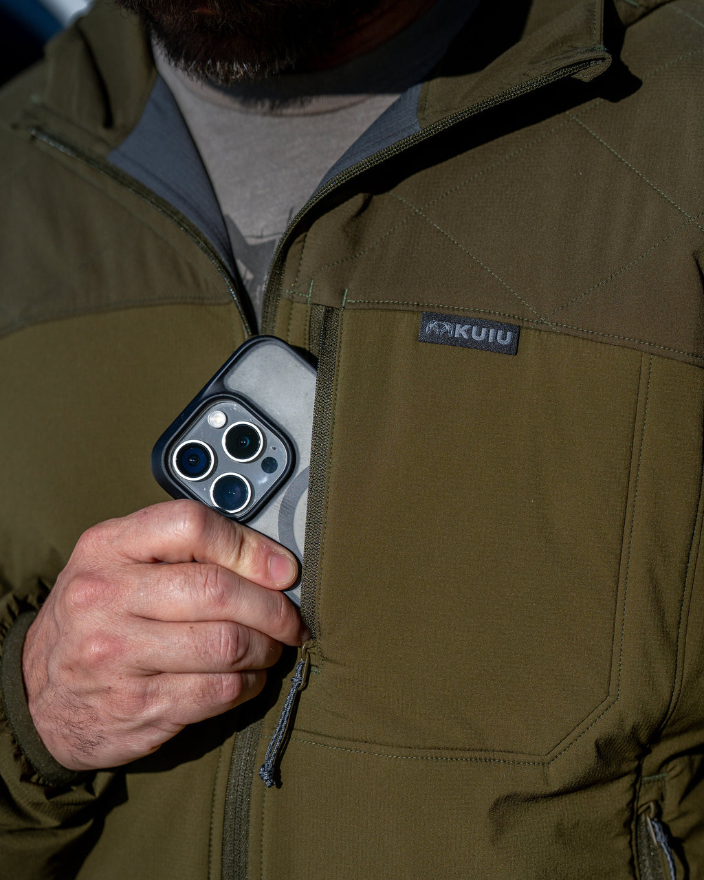 Close-up of a person wearing Transit Packable Hooded Jacket in Dark Army, stowing a smartphone in the chest pocket.