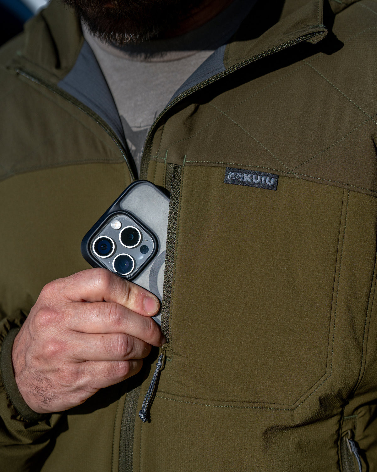 Close-up of a person wearing Transit Packable Hooded Jacket in Dark Army, stowing a smartphone in the chest pocket.