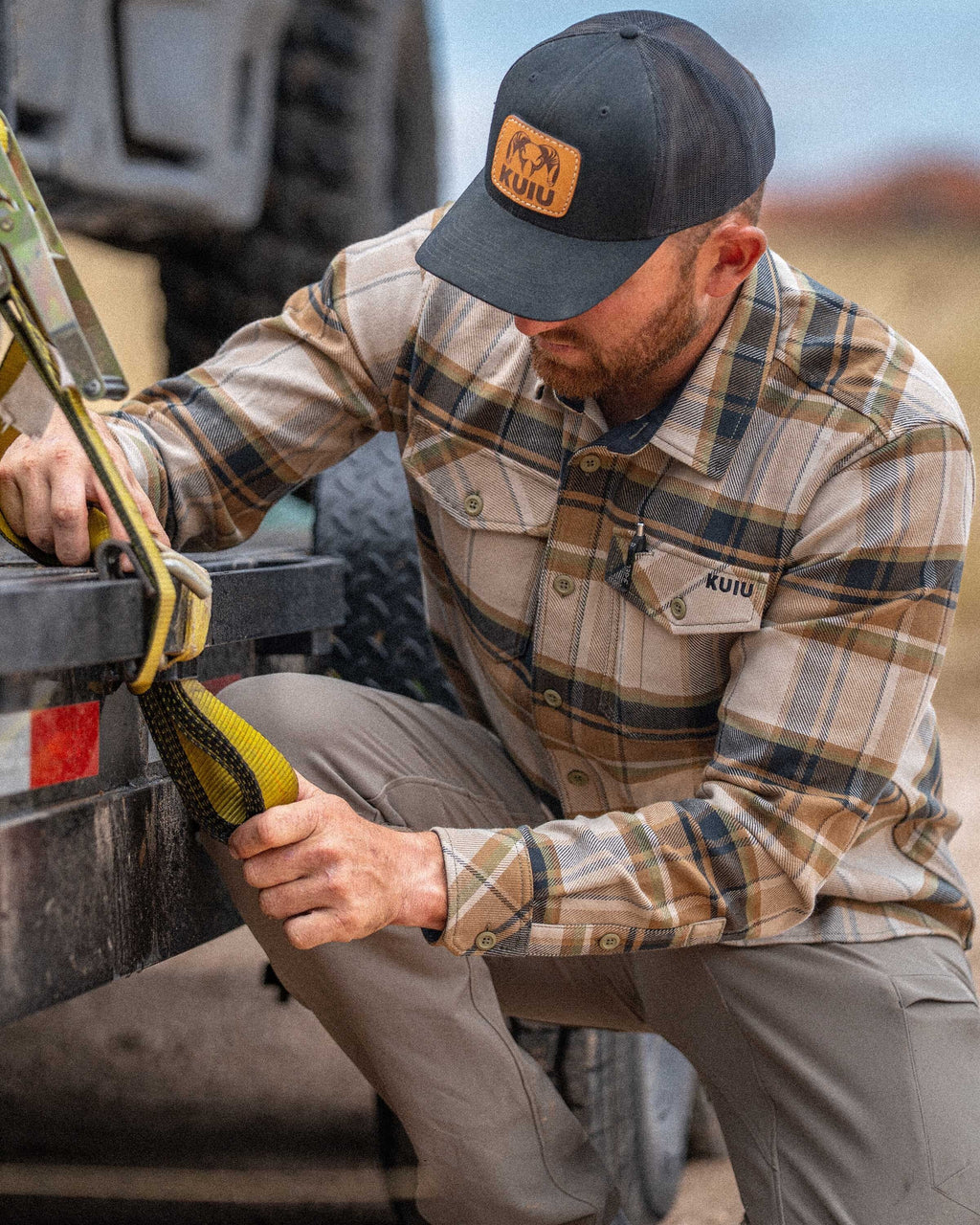 Man wearing Flannel Heavyweight Shirt in Valo Check Plaid kneels next to a hauler tightening a strap