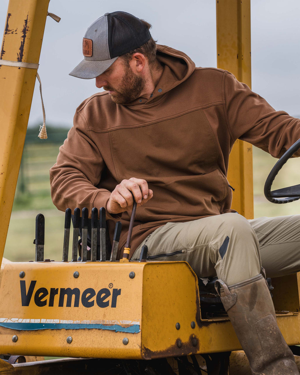 Man wearing Workgrade Utility Hoodie in Saddle Brown steering a yellow Vermeer construction machine