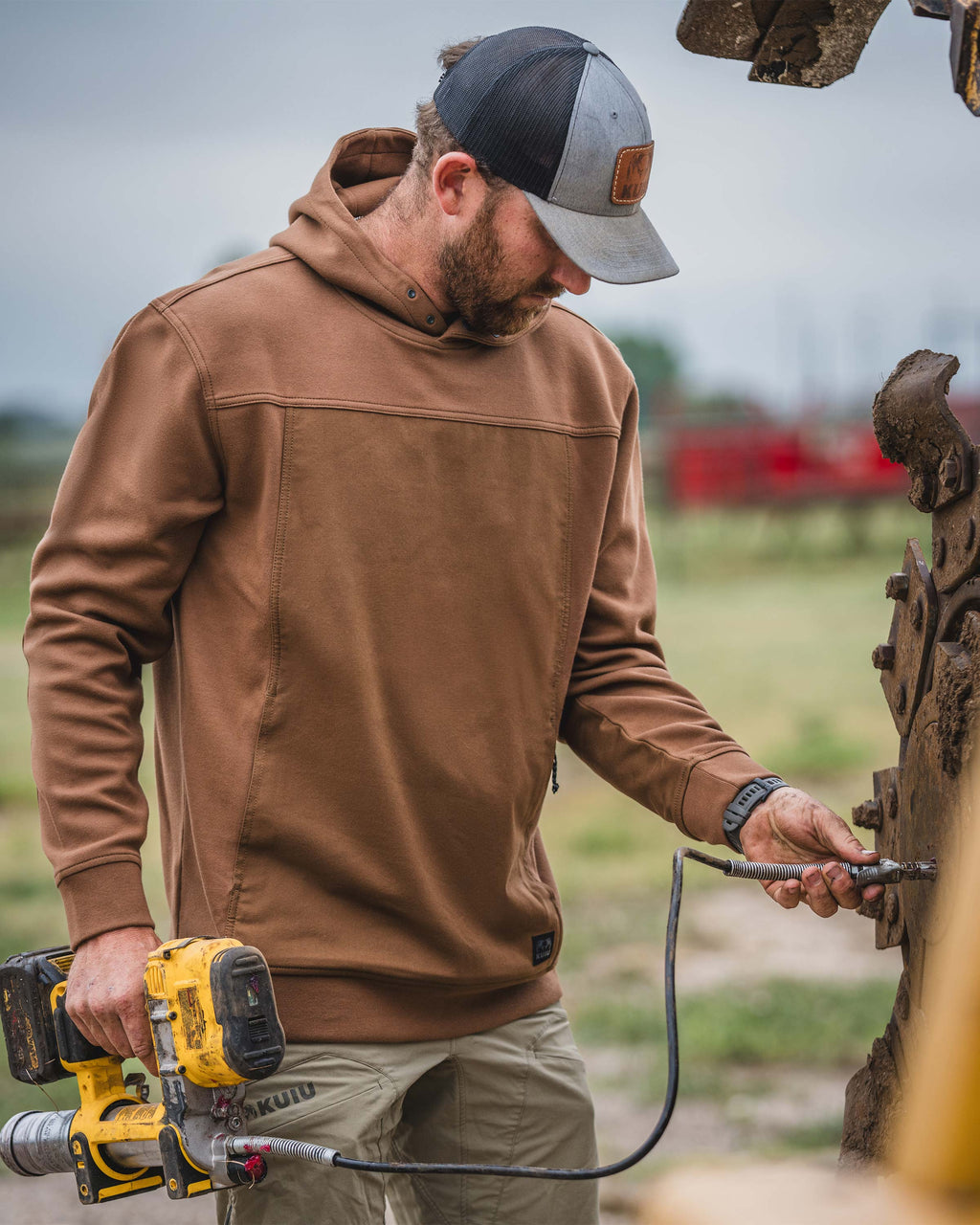 Man wearing Workgrade Utility Hoodie in Saddle Brown, holding a yellow power tool in the right hand while connecting a cable to a machinery with the left hand