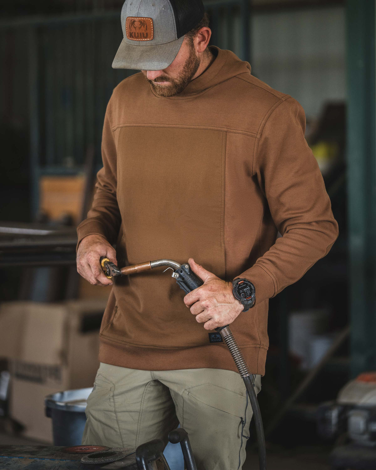 Man stands in a workshop, wearing Workgrade Utility Hoodie in Saddle Brown holding and inspecting a welding torch 
