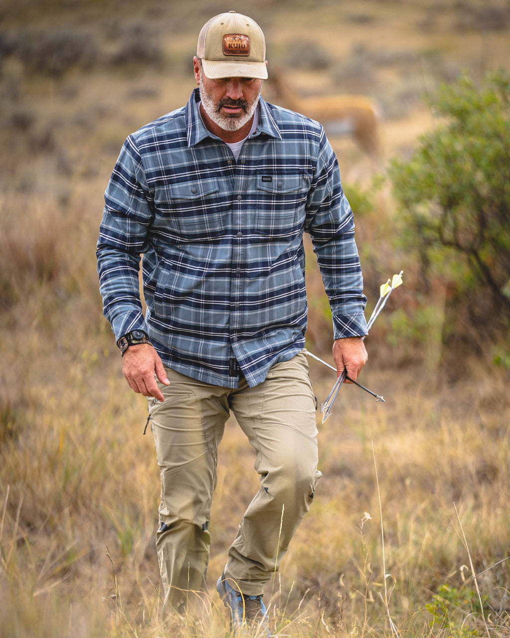 A man walking through a grassy outdoor field wearing KUIU Field Flannel Fleece in Steel Blue Framework, while holding a bow and arrow.